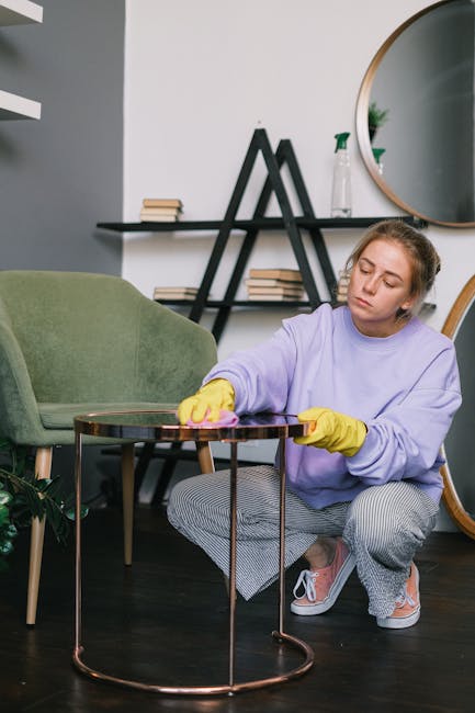 A person's hand holding a vacuum cleaner attachment, applying surface cleaning to a beige upholstered armchair seat with a perforated fabric. The armchair is situated in a well-lit room, and the clean